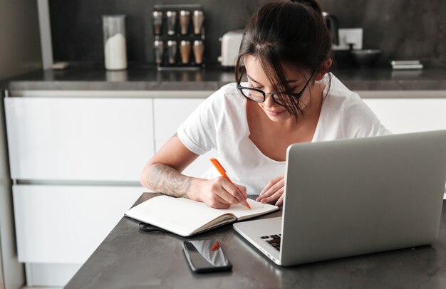 Jovem mulher séria concentrada que usa notas da escrita do laptop.