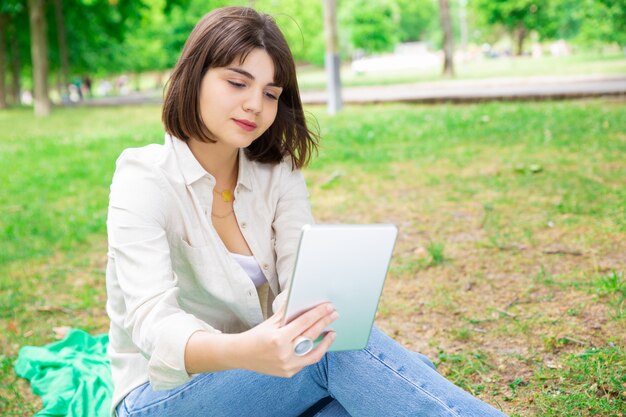 Jovem mulher séria que lê a notícia na tabuleta e que senta-se no gramado