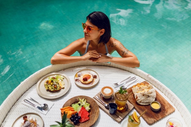 Jovem mulher sexy com tatuagem em fato de banho tomando café da manhã em uma piscina privada. Garota relaxante na piscina, bebendo café e comendo frutas. Prato de frutas, tigela de batidos à beira da piscina do hotel.