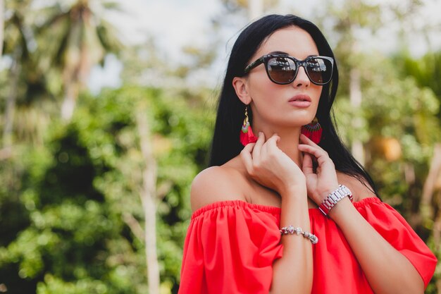 Jovem mulher sexy elegante com vestido vermelho de verão em pé no terraço de um hotel tropical, fundo de palmeiras, cabelo preto comprido, óculos de sol, brincos étnicos, óculos de sol, olhando para a frente, close-up