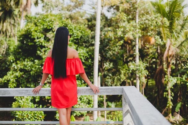 Jovem mulher sexy elegante com vestido vermelho de verão em pé no terraço de um hotel tropical, fundo de palmeiras, cabelo preto comprido, óculos de sol, brincos étnicos, óculos de sol, olhando para a frente