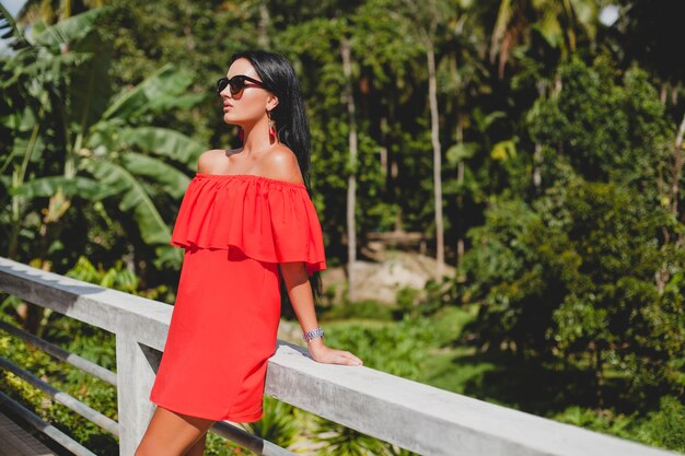 Jovem mulher sexy elegante com vestido vermelho de verão em pé no terraço de um hotel tropical, fundo de palmeiras, cabelo preto comprido, óculos de sol, brincos étnicos, óculos de sol, sorrindo