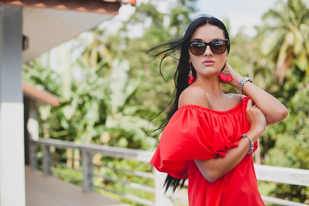 Jovem mulher sexy elegante com vestido vermelho de verão em pé no terraço de um hotel tropical, fundo de palmeiras, cabelo preto comprido, óculos de sol, brincos étnicos, óculos de sol, sorrindo