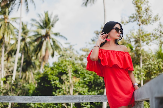 Jovem mulher sexy elegante com vestido vermelho de verão em pé no terraço de um hotel tropical, fundo de palmeiras, cabelo preto comprido, óculos de sol, brincos étnicos, óculos de sol, sorrindo
