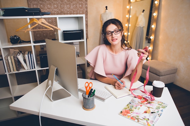 Jovem mulher sexy elegante em um vestido rosa de luxo, tendência de verão, estilo chique, estilista trabalhando no escritório no computador