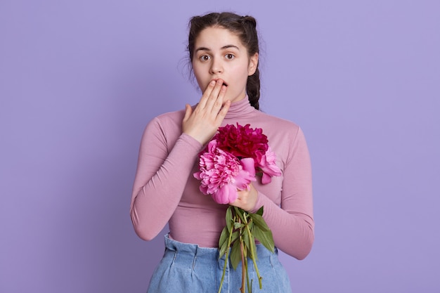 Jovem mulher sobre uma parede lilás, vestindo trajes casuais, dobrando flores nas mãos, ficando chocada, cobrindo a boca com a mão por engano, posando contra a parede lilás.
