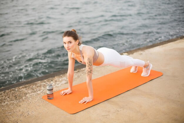 Foto grátis jovem mulher sonhadoramente olhando na câmera enquanto pratica ioga à beira-mar. menina bonita no top esportivo e leggings brancas fazendo exercícios esportivos no tapete de ioga laranja com vista para o mar no fundo