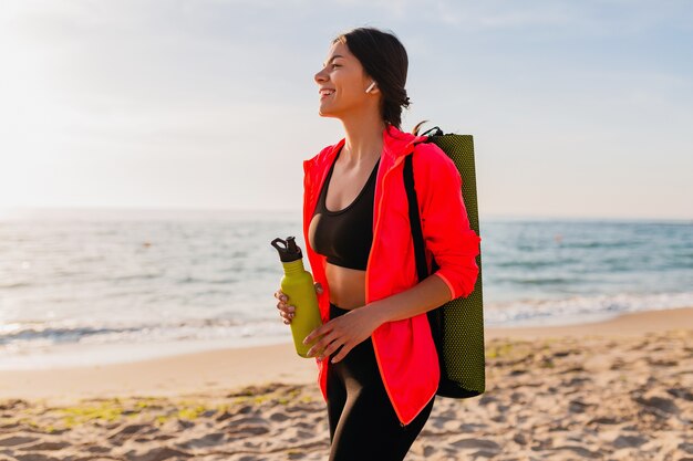 Jovem mulher sorridente atraente praticando esportes no nascer do sol da manhã na praia do mar segurando um tapete de ioga e uma garrafa de água, estilo de vida saudável, ouvindo música em fones de ouvido e vestindo uma jaqueta corta-vento rosa