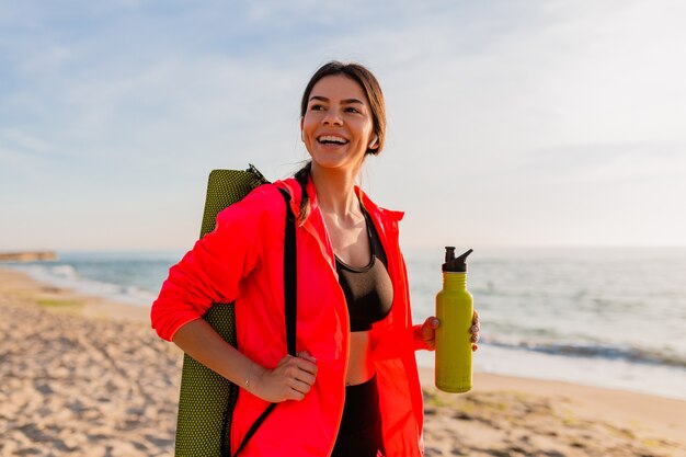 Jovem mulher sorridente atraente praticando esportes no nascer do sol da manhã na praia do mar segurando um tapete de ioga e uma garrafa de água, estilo de vida saudável, ouvindo música em fones de ouvido e vestindo uma jaqueta corta-vento rosa
