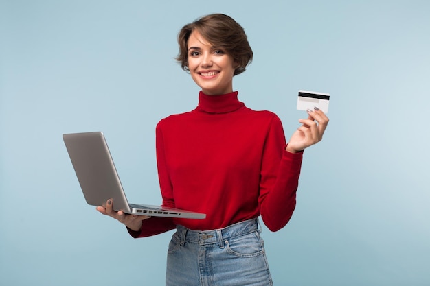 Jovem mulher sorridente com cabelo curto escuro na camisola vermelha e jeans segurando laptop e cartão de crédito nas mãos enquanto alegremente olhando na câmera sobre fundo azul