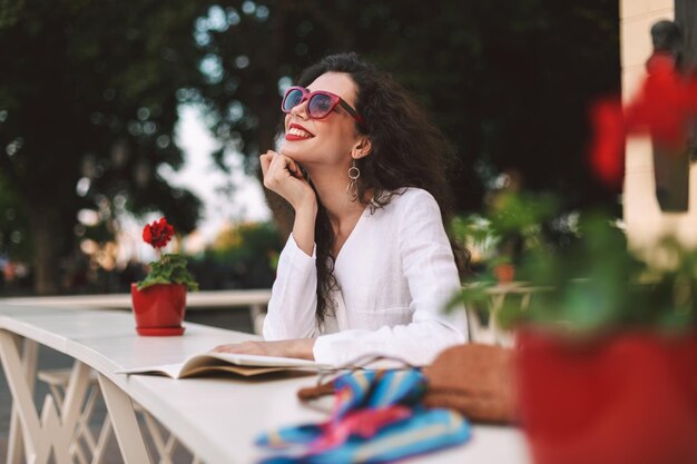 Jovem mulher sorridente com cabelo escuro encaracolado em óculos de sol em pé com o bloco de notas e sonhadoramente olhando de lado enquanto passa o tempo no terraço de verão do café