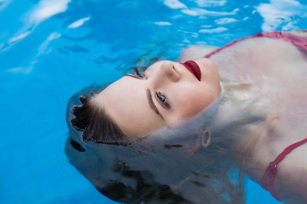 Jovem mulher sorridente de biquíni relaxa relaxante, nadando de costas em águas claras na piscina. Garota gostosa em trajes de banho encontra-se na água no sol nas férias de verão.