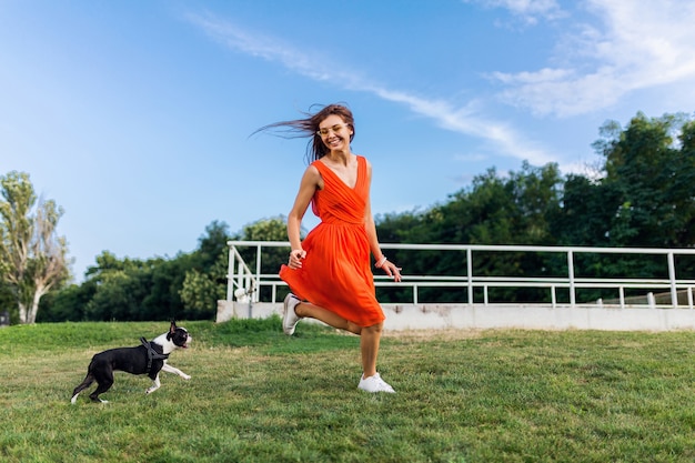 Jovem mulher sorridente feliz com vestido laranja se divertindo, brincando, correndo com o cachorro no parque, estilo verão, clima alegre