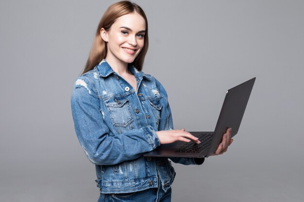 Jovem mulher sorridente feliz em roupas casuais, segurando o laptop e enviar e-mail para sua melhor amiga isolada na parede cinza