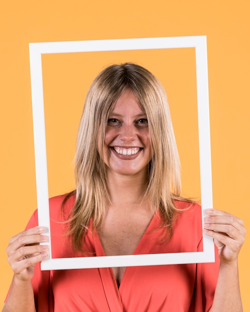 Jovem mulher sorridente segurando a moldura de foto de borda branca na frente de seu rosto