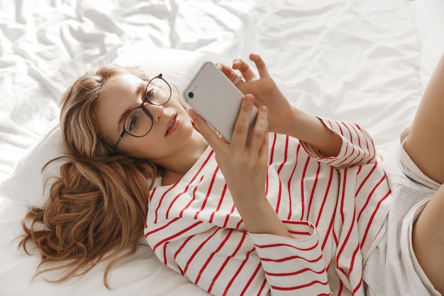 Jovem mulher sorridente, sentada na cama em casa e mandando mensagens de texto com seu smartphone.