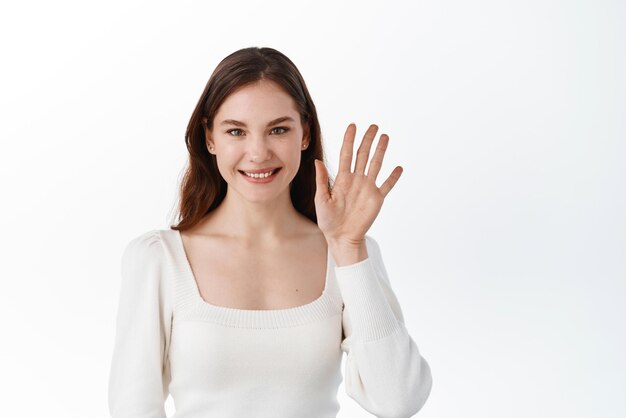 Jovem mulher sorrindo e saudando acenando com a mão para dizer olá, cumprimentando os amigos, diga oi e dê boas-vindas ao gesto de adeus em pé contra o fundo branco