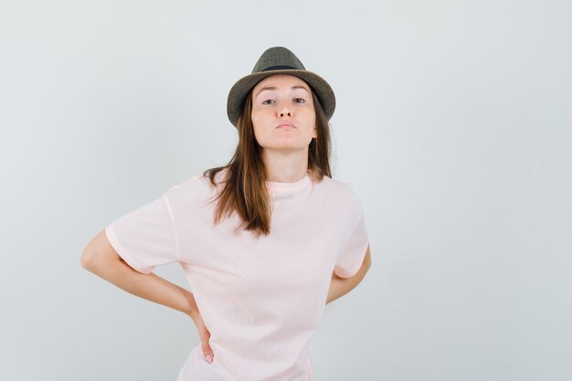 Jovem mulher tendo dor nas costas em t-shirt rosa, chapéu e parecendo exausta, vista frontal.