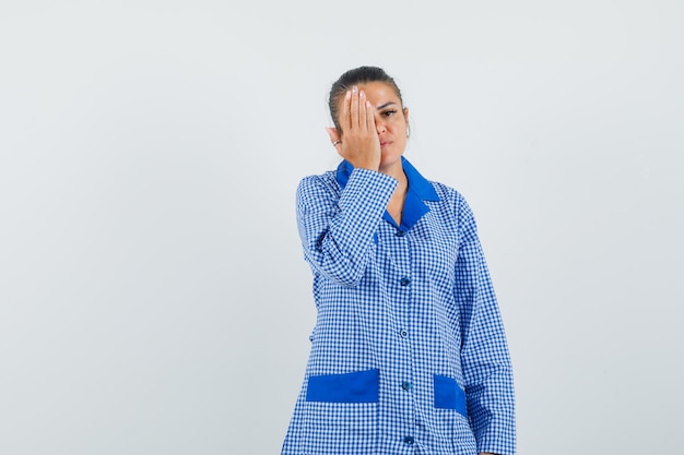 Jovem mulher tentando cobrir o olho com a mão na camisa do pijama guingão azul e olhando sério, vista frontal.