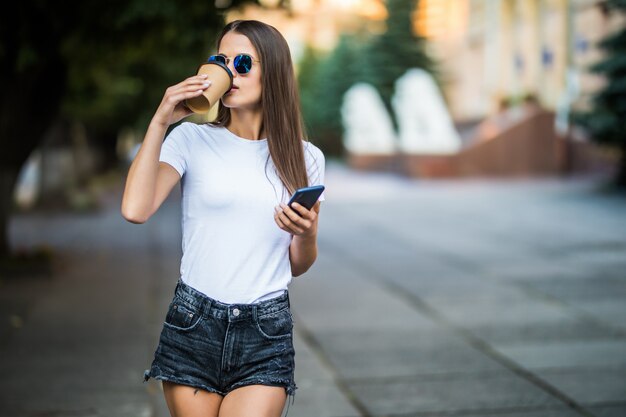 Jovem mulher tomando um café e usando o smartphone na rua