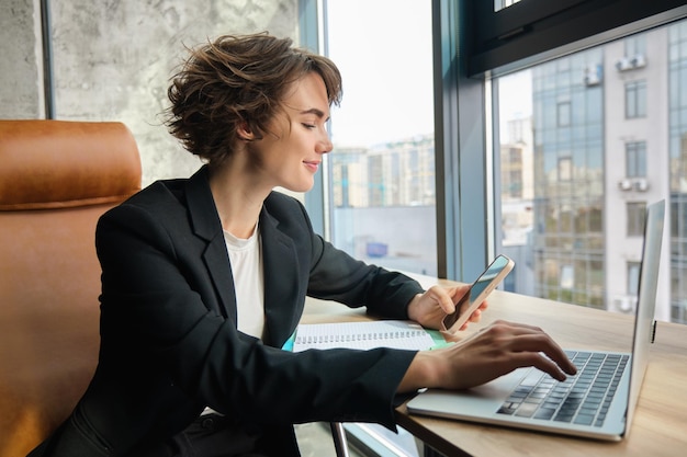 Jovem mulher trabalhadora no escritório, sentada em frente ao computador e usando smartphone, vestindo terno