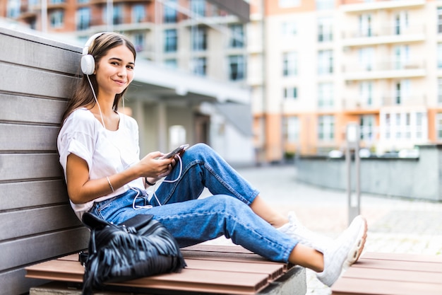 Jovem mulher usando telefone celular ouve música enquanto está sentado no banco de um parque