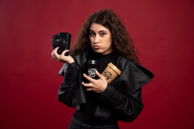 Jovem mulher vestida de preto, segurando copos e uma câmera.