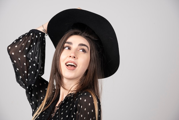 Jovem mulher vestida de preto, segurando o chapéu no topo da cabeça.