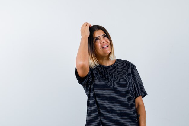 Jovem mulher vestida de preto, segurando uma mão na cabeça, pensando em algo e olhando pensativa, vista frontal.