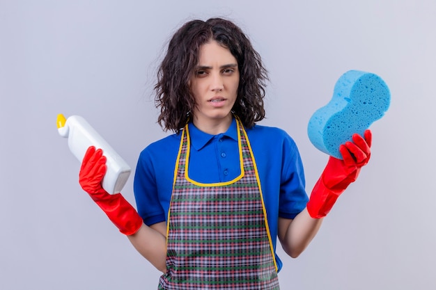 Foto grátis jovem mulher vestindo avental e luvas de borracha, segurando a esponja e produtos de limpeza com expressão suspeita sobre parede branca