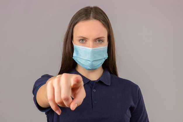 Jovem mulher vestindo camisa polo azul em máscara médica protetora, apontando o dedo para a câmera com cara séria em pé sobre fundo cinzento claro