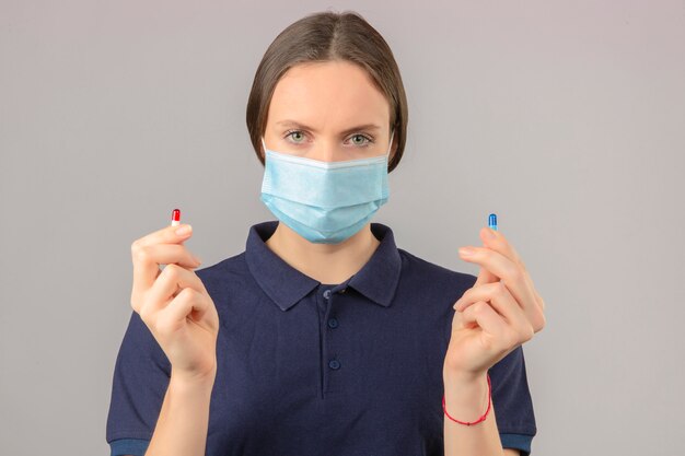 Jovem mulher vestindo camisa polo azul em máscara médica protetora segurando comprimidos na mão, olhando para a câmera com cara séria sobre fundo cinza claro