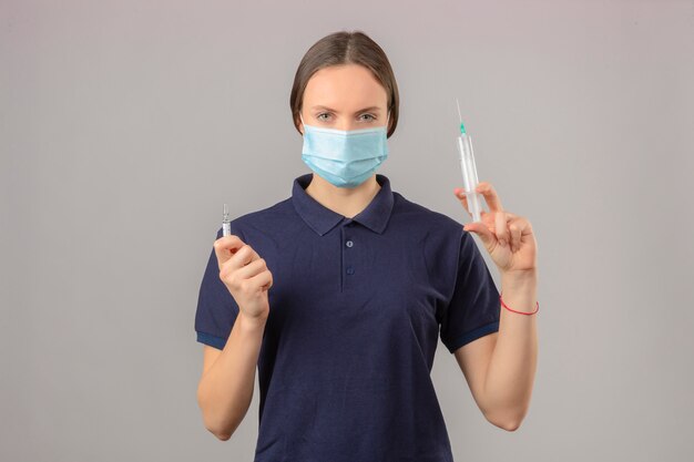 Jovem mulher vestindo camisa polo azul em máscara médica protetora, segurando o frasco de vacina seringa e medicamento, olhando para a câmera com cara séria em pé sobre fundo cinzento isolado