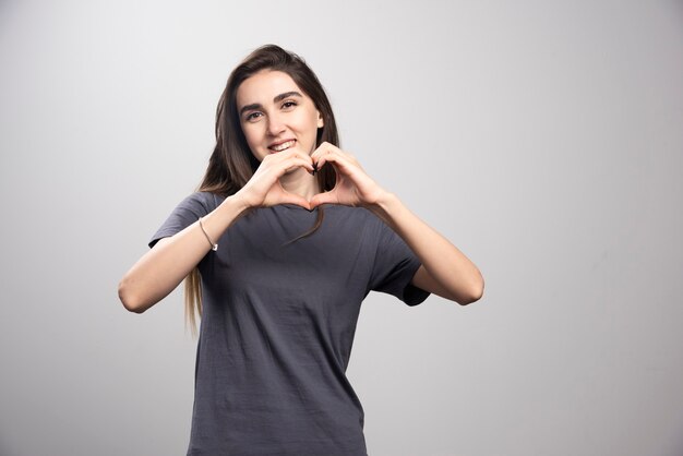 Jovem mulher vestindo camiseta cinza sobre fundo cinza, fazendo a forma do símbolo do coração com as mãos.