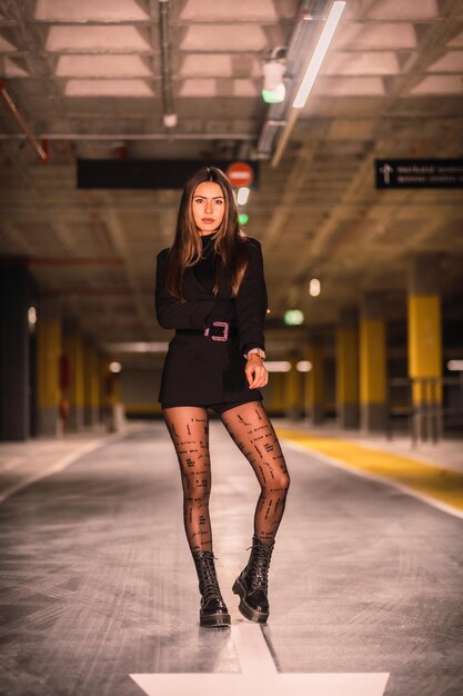 Jovem mulher vestindo uma jaqueta preta com shorts e botas e posando em um estacionamento subterrâneo