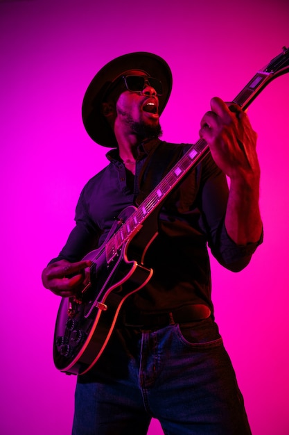 Foto grátis jovem músico afro-americano tocando guitarra como uma estrela do rock em fundo gradiente rosa-púrpura em luz de néon.