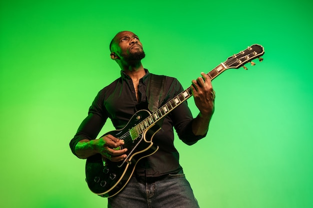 Jovem músico afro-americano tocando guitarra como uma estrela do rock em fundo gradiente verde-amarelo. Conceito de música, hobby, festival, ao ar livre. Cara alegre improvisando, cantando música.