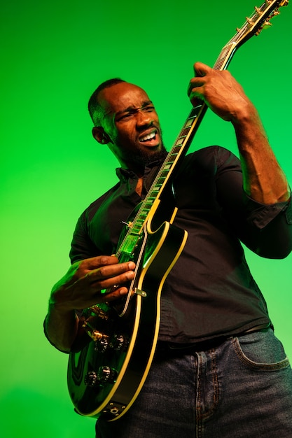 Jovem músico afro-americano tocando guitarra como uma estrela do rock em fundo gradiente verde-amarelo. Conceito de música, hobby, festival, ao ar livre. Cara alegre improvisando, cantando música.