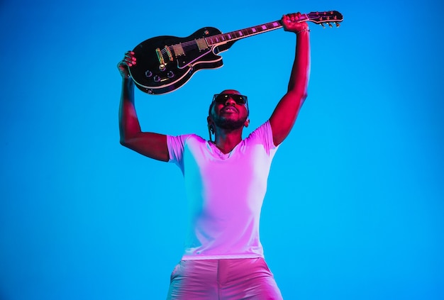 Jovem músico afro-americano tocando violão como uma estrela do rock na parede azul em luz de néon.