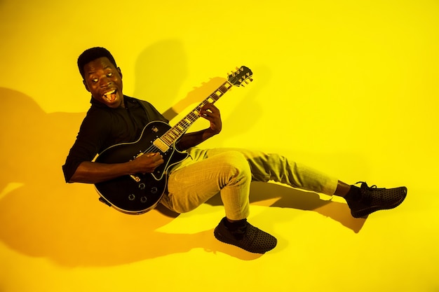 Foto grátis jovem músico afro-americano tocando violão como uma estrela do rock sobre fundo amarelo em luz de néon.