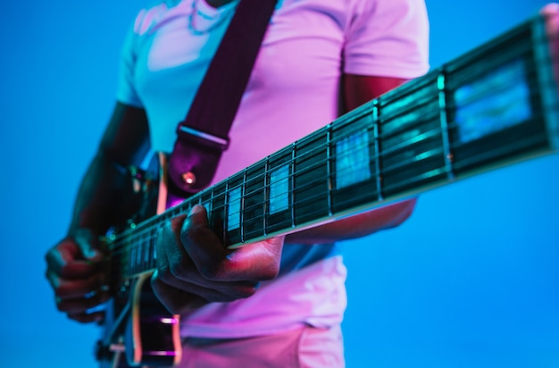 Jovem músico afro-americano tocando violão como uma estrela do rock sobre fundo azul do estúdio em luz de néon. Conceito de música, hobby. Cara alegre improvisando. Retrato retro colorido.