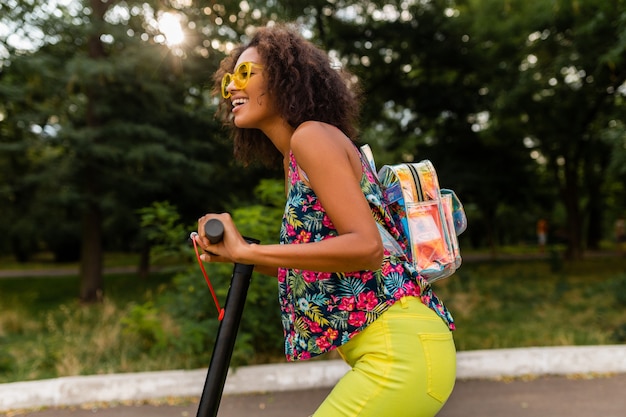 Jovem negra elegante se divertindo no parque, andando de scooter elétrica no estilo da moda de verão, roupa colorida hipster, usando mochila e óculos de sol amarelos