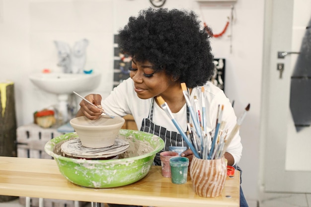 Foto grátis jovem negra pintando panela de barro no estúdio de arte artesã pintando tigela de cerâmica na bancada garota vestindo um avental azul