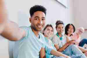 Foto grátis jovem negro bonito com penteado encaracolado fazendo selfie com amigos e sorrindo. retrato interno de alunos rindo alegres, se divertindo depois de lessong e tirando foto.