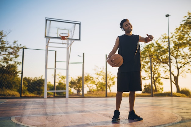 Jovem negro sorridente e feliz praticando esportes, jogando basquete ao nascer do sol, ouvindo música em fones de ouvido, estilo de vida ativo, manhã de verão