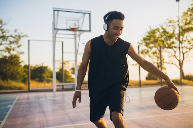 Jovem negro sorridente e feliz praticando esportes, jogando basquete ao nascer do sol, ouvindo música em fones de ouvido, estilo de vida ativo, manhã de verão