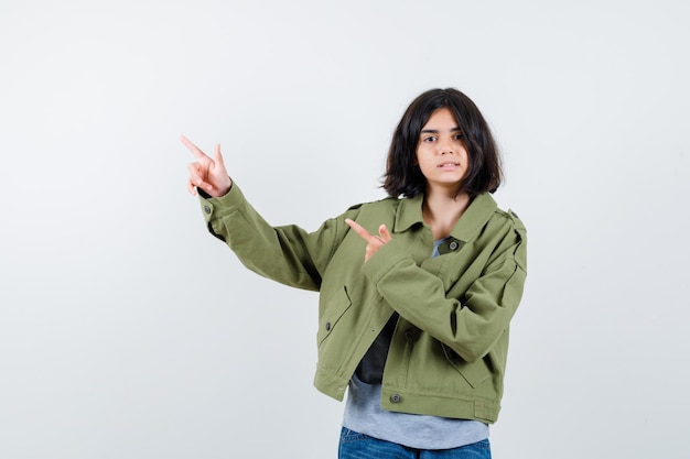 Foto grátis jovem num suéter cinza, jaqueta cáqui, calça jeans apontando para a esquerda com o dedo indicador e olhando séria, vista frontal.