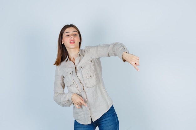 Jovem numa camisa bege, jeans apontando para baixo com o dedo indicador e olhando com foco, vista frontal.