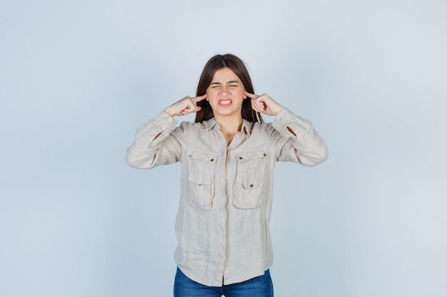 Foto grátis jovem numa camisa bege, jeans tapando as orelhas com o dedo indicador e parecendo agitada, vista frontal.