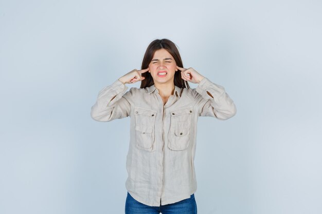 Jovem numa camisa bege, jeans tapando as orelhas com o dedo indicador e parecendo agitada, vista frontal.